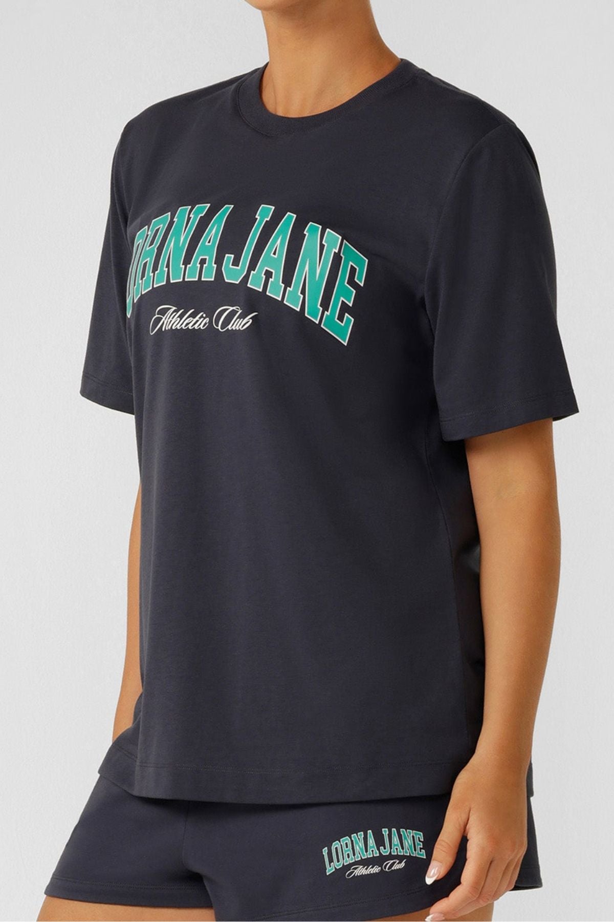 Wearing a relaxed fit Lorna Jane Original Sport Relaxed T-Shirt with matching sporty shorts, both featuring logos. The tee displays "Lorna Jane Athletic Club" in bold teal and white; the person's head is out of frame.