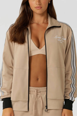 A woman wears the Lorna Jane Retro Track Zip Through Jacket with an athletic fit, black sleeve stripes, chest embroidery, and zip pockets, styled over a beige sports bra and joggers.