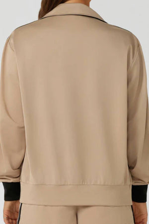 A person is shown from the back wearing the Lorna Jane Retro Track Zip Through Jacket in beige with black cuffs and matching pants. The jacket has a relaxed, athletic fit and a simple retro design.