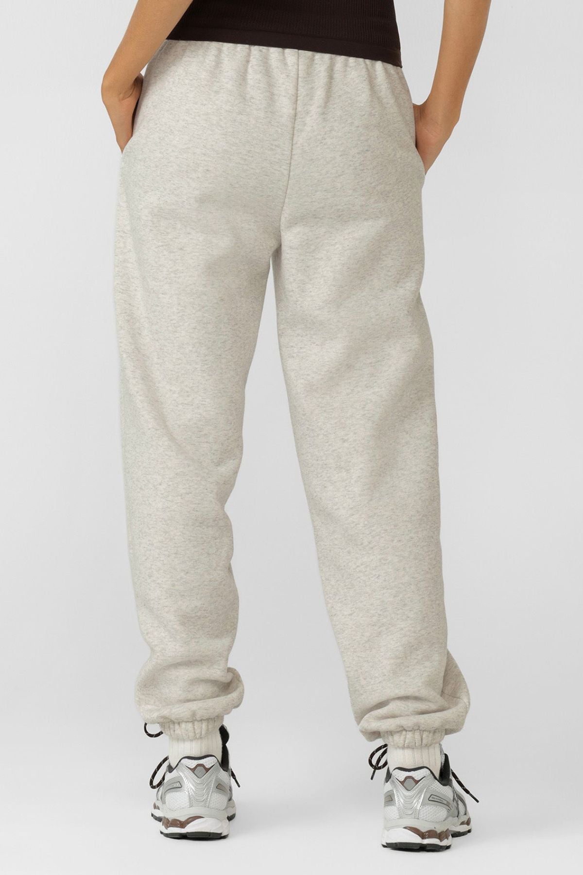 Someone in relaxed fit Lorna Jane Sport Mode Sweatpants made from cotton blend fleece, white sneakers, and a dark top stands facing away with hands in pockets against a plain light background.