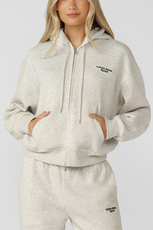 A woman stands against a plain background wearing the Lorna Jane Sport Mode Zip Through Hoodie in light grey cotton-blend fleece, featuring "Lorna Jane Sport" logos and an adjustable drawcord hood, with her hands in her pockets.