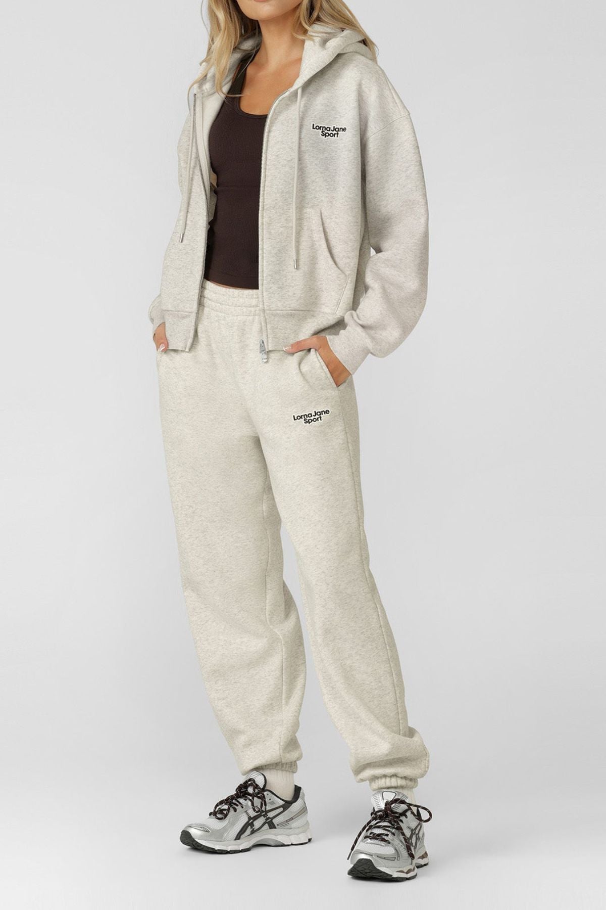 A woman wearing a Lorna Jane Sport Mode Zip Through Hoodie in light gray with black text, matching sweatpants, a dark brown tank top, and gray sneakers stands against a plain light background.