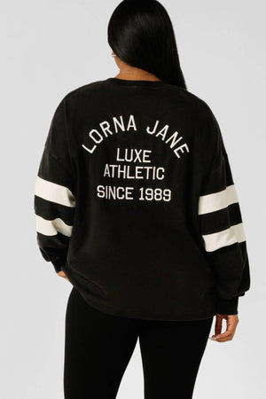 A person with long dark hair wears the Lorna Jane Throwback Oversized Lightweight Sweat, featuring white sleeve stripes and "Lorna Jane Luxe Athletic Since 1989" printed on the back.