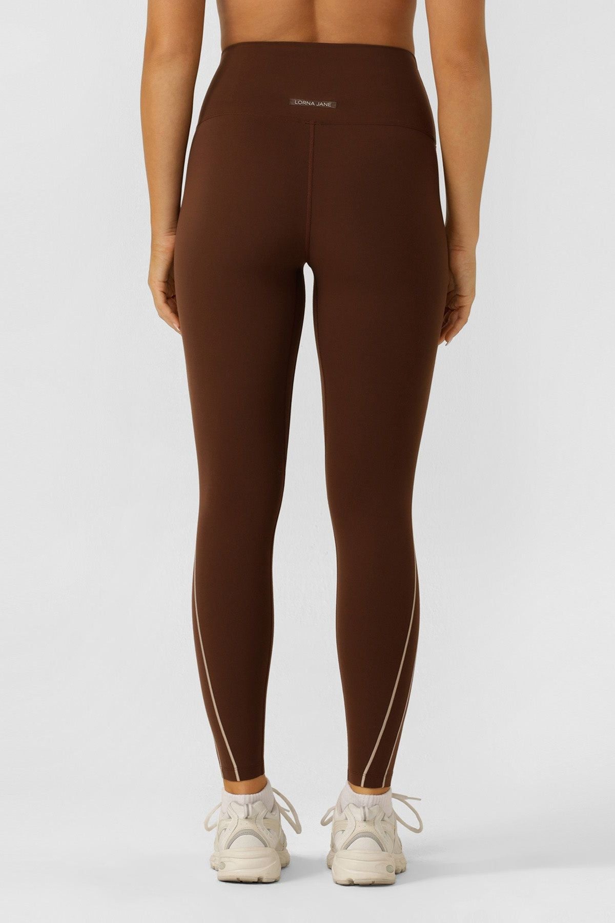 Wearing Lorna Jane Total Bliss Ankle Biter Leggings with a high-rise waistband and subtle seams, a person stands with their back to the camera. The brown 4-way stretch leggings are paired with white sneakers against a plain light background.