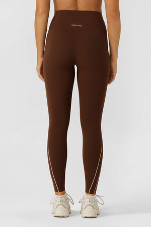 Wearing Lorna Jane Total Bliss Ankle Biter Leggings with a high-rise waistband and subtle seams, a person stands with their back to the camera. The brown 4-way stretch leggings are paired with white sneakers against a plain light background.
