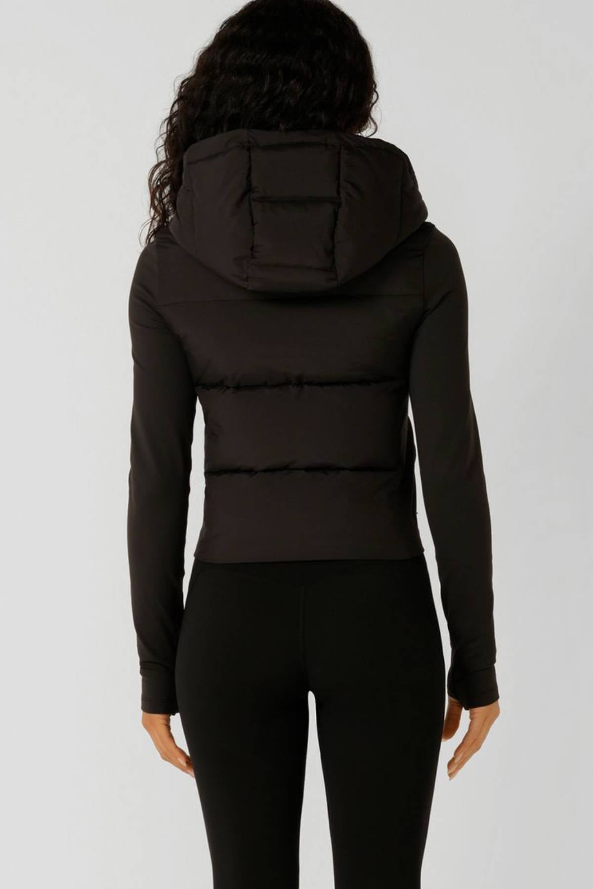 A person with long, curly hair is seen from behind wearing the Lorna Jane Warm Up Thermal Combo Puffer with an adjustable hood and black fitted pants against a plain light background.
