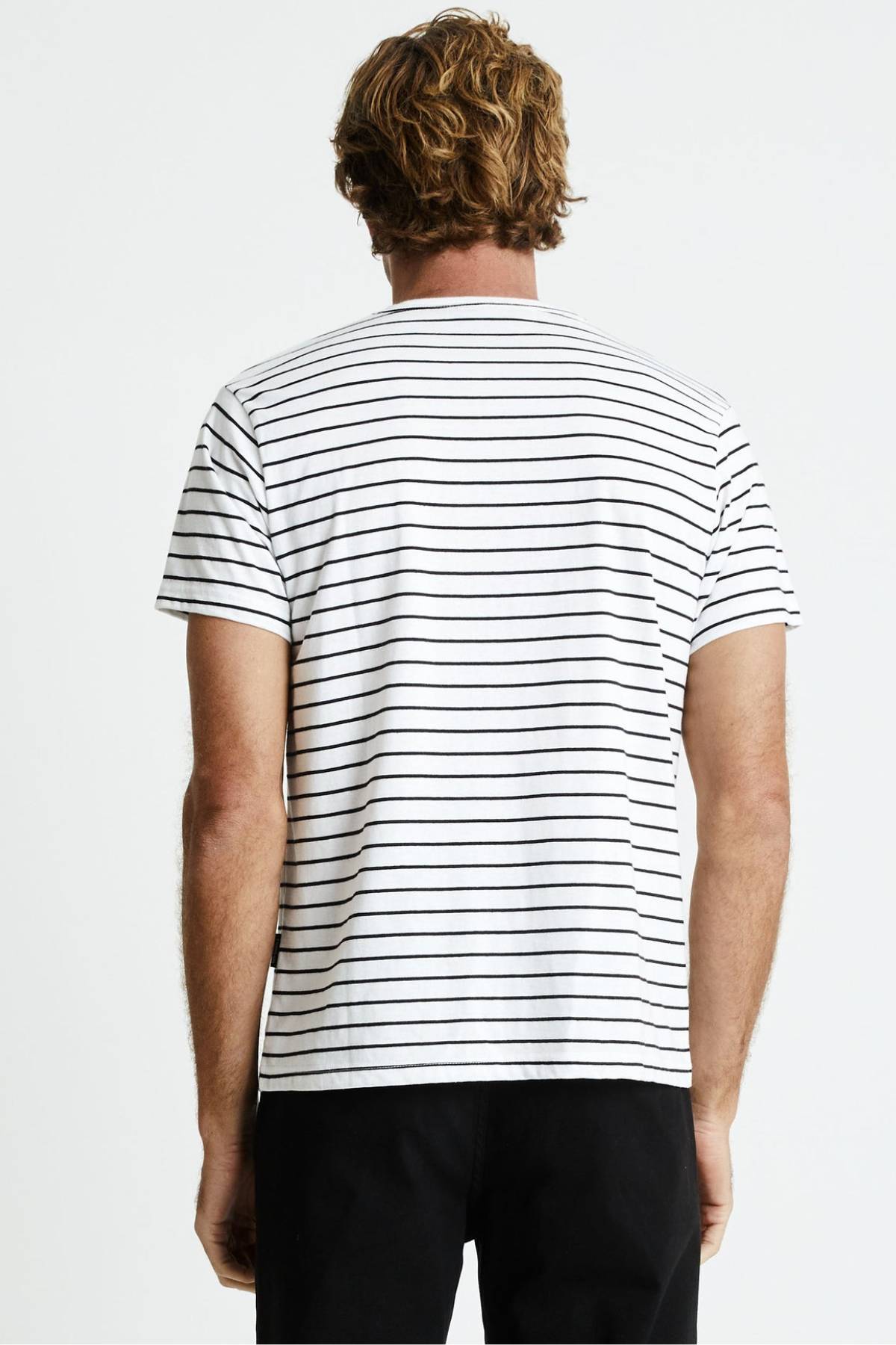 A person with wavy, light brown hair is standing with their back to the camera. They are wearing a Mr Simple Breton Stripe Tee made from 100% organic cotton, paired with black pants. The short-sleeved white t-shirt features horizontal black stripes, and the background is plain white.