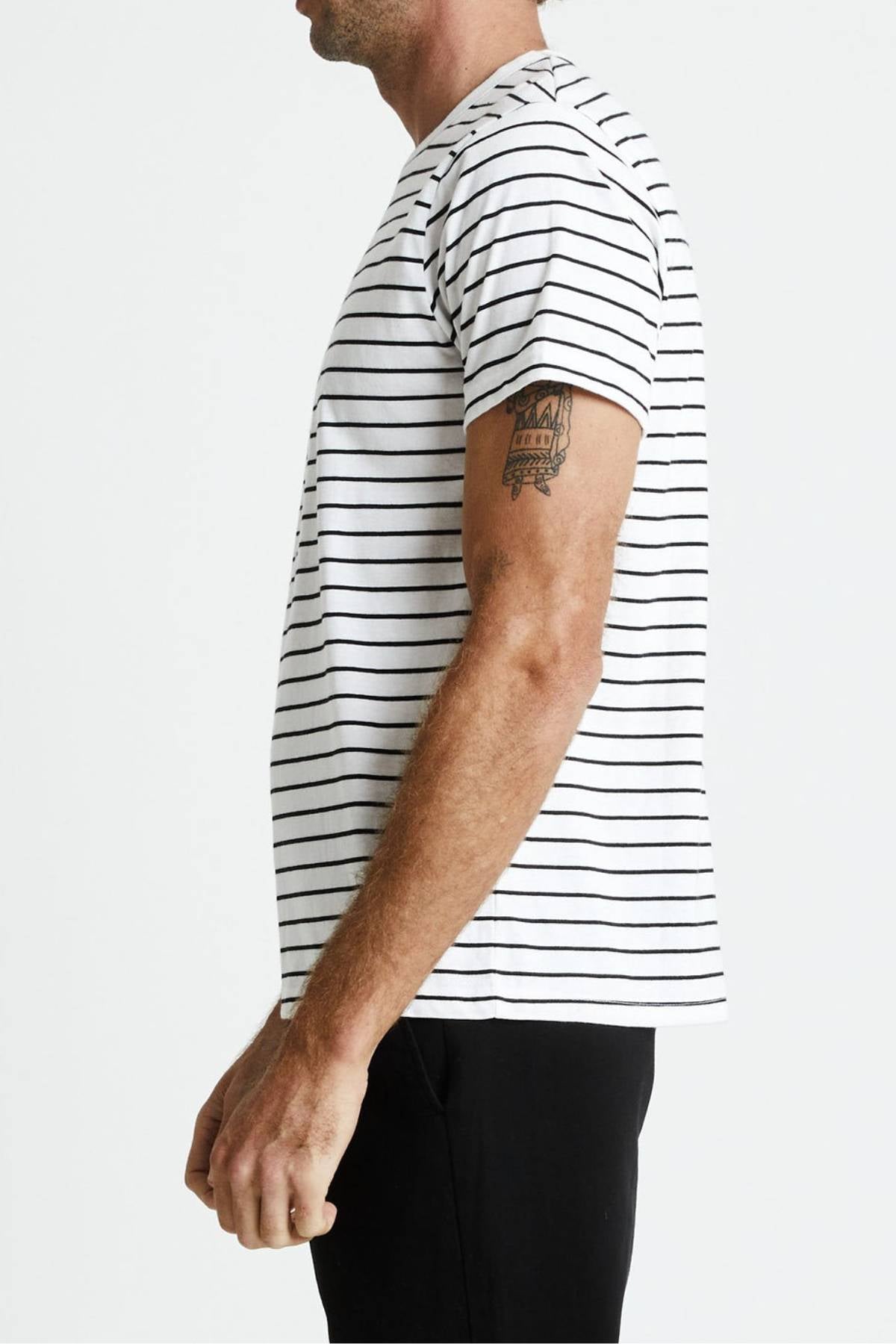 Side view of a person wearing the Mr Simple Breton Stripe Tee with black pants. The 100% organic cotton white t-shirt boasts black horizontal stripes. The individual, displaying a tattoo on their upper arm, stands against a plain background.
