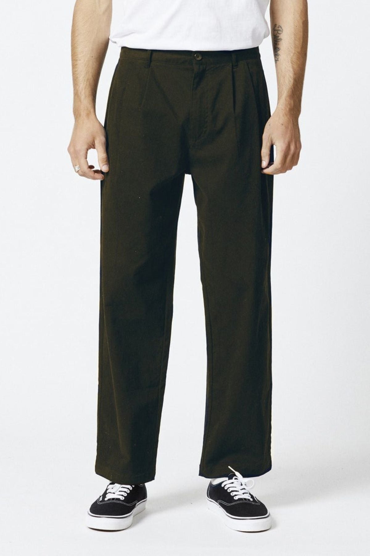 A person wears the Mr Simple Brooklyn Pleated Pant in olive green with a relaxed fit, paired with a white t-shirt and black sneakers, standing against a plain white background. The image is cropped above the shoulders to highlight the outfit.