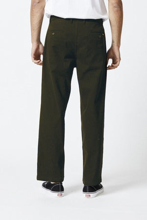 A person wearing the Mr Simple Brooklyn Pleated Pant in dark green, paired with a white shirt and black sneakers, stands with their back to the camera against a plain white background.