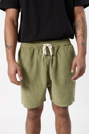 A person is shown from the shoulders down, wearing a black shirt and the Mr Simple Burbank Cord Short in olive green with a white drawstring. The person's arms, adorned with tattoos, are visible as they stand against a plain white background.