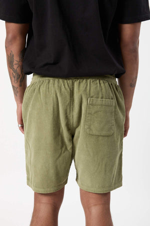 A person is shown from the back wearing a black t-shirt and Mr Simple Burbank Cord Shorts in light green, which feature an elastic waistband and a single back pocket on the right side. The person's tattooed arms are visible.