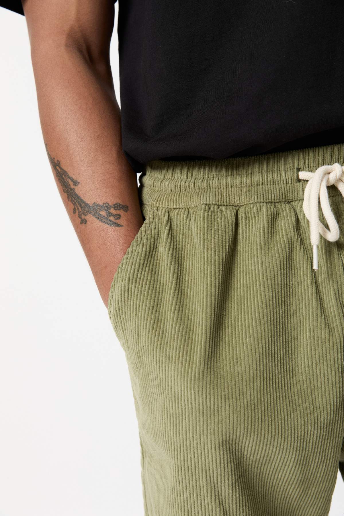 Close-up of a person wearing Mr Simple Burbank Cord Shorts in olive-green with a white drawstring. The person has tanned skin and a tattoo of branches on their left forearm. They are also wearing a black shirt, and their left hand is in the pocket of the shorts.