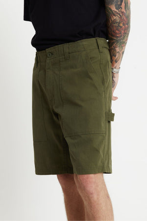 Side view of a person wearing the relaxed fit olive green Mr Simple Carpenter Walk Short paired with a black shirt. The person's arm is visible, showcasing a multicolored tattoo sleeve and adorned with a light-colored bracelet. The herringbone cotton shorts feature visible pockets and a loop detail on the side.