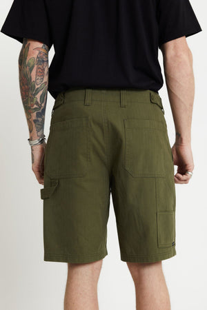 A person wearing a black shirt and green Mr Simple Carpenter Walk Short is shown from the back. One arm is tattooed with a floral design, and the person is also wearing a silver bracelet. The relaxed fit shorts feature several pockets and a loop on one side, ideal for both style and utility.