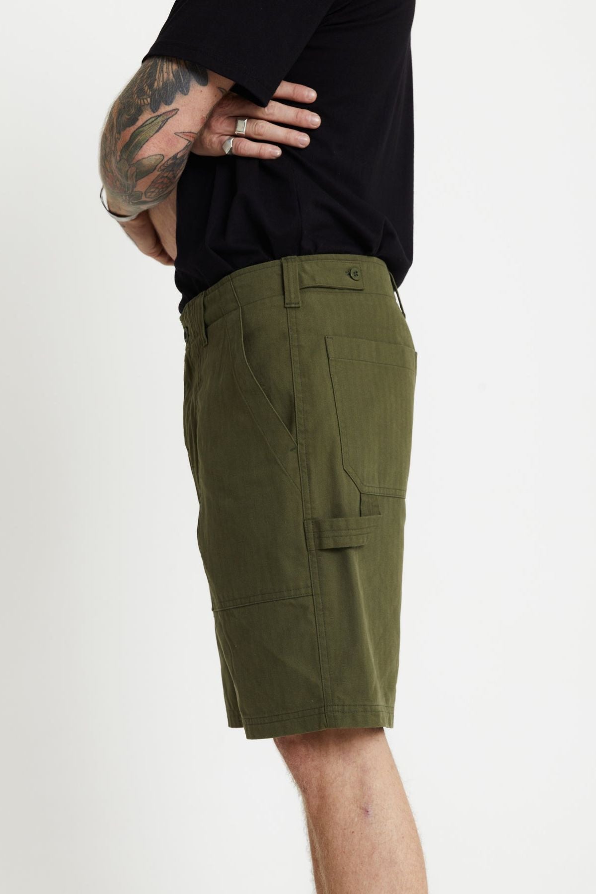 Side profile of a person wearing a black t-shirt and relaxed fit shorts. They have several tattoos on their arm and are standing against a plain white background. The Mr Simple Carpenter Walk Short in olive green adds a rugged yet stylish touch to their look.