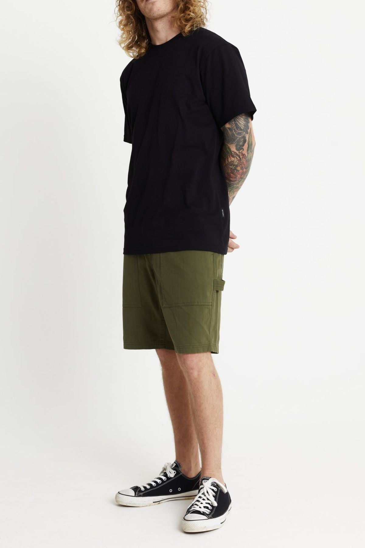 A person with long curly hair stands against a plain white background. They are wearing a black T-shirt, Mr Simple Carpenter Walk Shorts, and black-and-white sneakers. Their hands are positioned behind their back, and they have tattoos visible on their forearms.