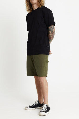 A person with long curly hair stands against a plain white background. They are wearing a black T-shirt, Mr Simple Carpenter Walk Shorts, and black-and-white sneakers. Their hands are positioned behind their back, and they have tattoos visible on their forearms.