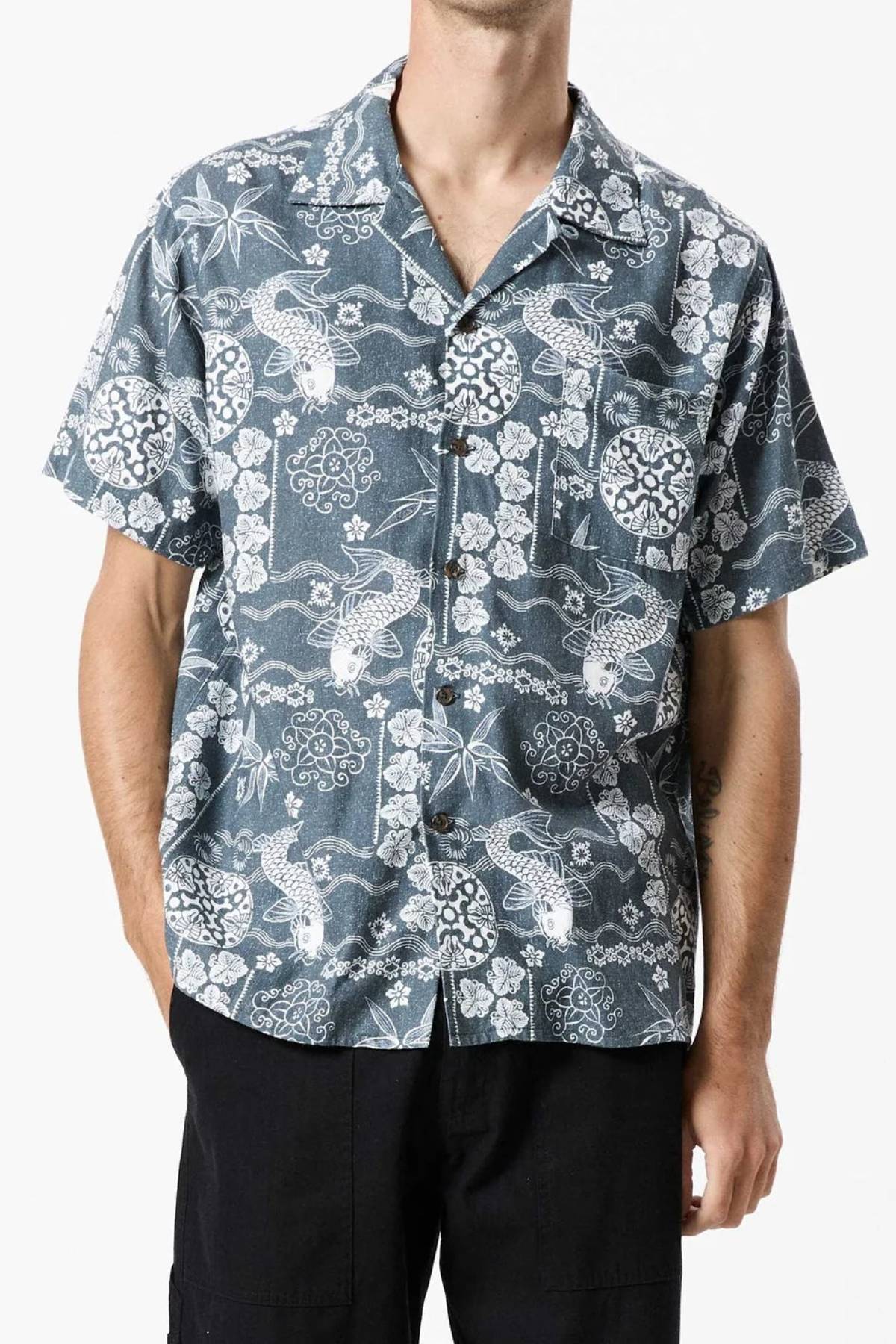 A person is wearing the Mr Simple Koi Bowler Short Sleeve Shirt, which features a blue and white floral and bird pattern. The individual is shown from the shoulders to the thighs, and the shirt has a relaxed, casual fit. The background is plain white.