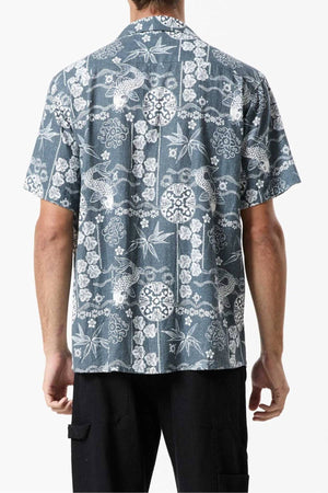 A person is wearing the Mr Simple Koi Bowler Short Sleeve Shirt, which boasts a vibrant print featuring fish, leaves, and geometric patterns in white against a dark blue background. The person is standing with their back facing the camera, showcasing the shirt’s intricate design.
