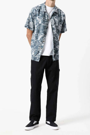 A person is standing against a plain white background, wearing the Mr Simple Koi Bowler Short Sleeve Shirt over a white T-shirt, black pants, and black slip-on shoes with white soles. The person's hands are behind their back, and their head is not visible in the image.