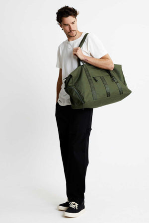A man in a white t-shirt and black pants carries a Mr Simple Nelson Duffle over his shoulder while wearing black sneakers with white soles. The plain white background accentuates the quality of the goods he holds.