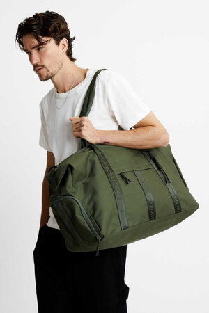 A man with short brown hair and a thin beard stands against a plain white background, wearing a white t-shirt and black pants while carrying a large olive-green Mr Simple Nelson Duffle. The bag features black straps and zippers, highlighting its QUALITY GOODS craftsmanship.