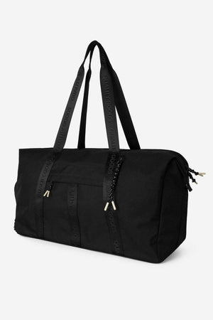 The Mr Simple Nelson Duffle is a large black bag with double shoulder straps, zipper closures, and "QUALITY GOODS" on the straps. It has a minimalist rectangular shape and a trolley sleeve for convenient travel.