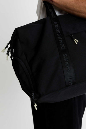 A person carries the Mr Simple Nelson Duffle, a large black bag with visible zippers, white pull tabs, “Quality Goods” on the strap, and a trolley sleeve—styled with a white top and black pants.