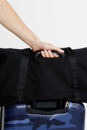 A person’s hand holds a dark blue camouflage suitcase with the Mr Simple Nelson Duffle in black secured on top via its trolley sleeve and a strap labeled "QUALITY GOODS.