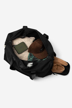 The Mr Simple Nelson Duffle, open on a white background, features a handy trolley sleeve and contains a knit sweater, green pouch, brown corduroy cap, folded clothes, and tan sandals packed neatly inside.