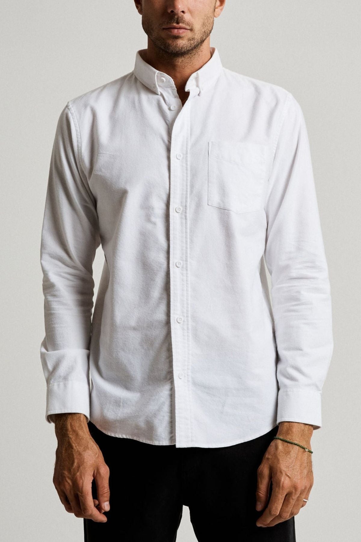 A man models the Mr Simple Oxford Shirt, a slim-fit white 100% cotton shirt with a front pocket, standing against a plain light gray background. His relaxed hands are at his sides, and only his upper body is shown.
