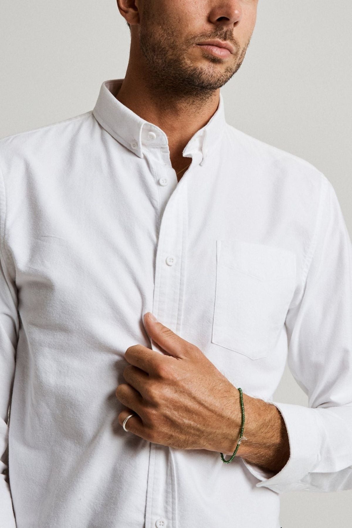 A man adjusts his Mr Simple Oxford Shirt—slim-fit and crafted from 100% cotton—in front of a plain background. He wears a silver ring and green bracelet on his left hand, highlighting the shirt’s comfort and style.