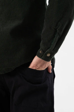 A person wears the Mr Simple Quilted Cord Jacket in dark green with black pants, standing with their right hand in their pocket, shown from the waist down against a plain background.