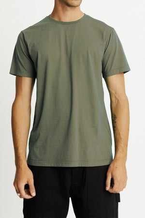A person wearing a Mr Simple Reginald Organic Tee in plain green and black pants stands against a light background. The person's face is not visible, and tattoos are partially visible on their left forearm.