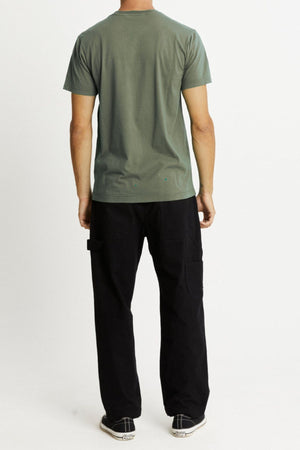 A man is standing with his back to the camera, wearing a Mr Simple Reginald Organic Tee in green, black cargo pants, and black and white sneakers. The background is plain and light-colored.
