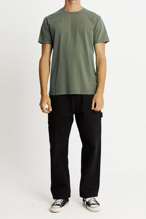 A person stands against a plain light background, wearing the Mr Simple Reginald Organic Tee in green, paired with loose-fitting black pants and black sneakers with white soles. The person's arms are relaxed at their sides, and the top of their head is not visible.