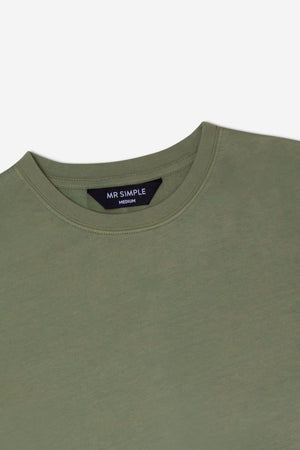 A close-up image of the upper part of the Mr Simple Reginald Organic Tee in olive green. The shirt features a round neckline and a tag at the back of the collar that reads "MR SIMPLE Medium" in white letters on a black background.