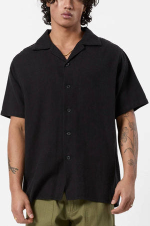 Wearing the Mr Simple Waffle Short Sleeve Shirt in black, 100% cotton with a collar, paired with olive green pants and showing forearm tattoos, someone stands against a plain light background—ideal for any casual wardrobe.