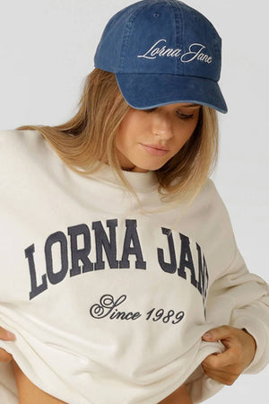 A person wearing a blue Lorna Jane Heritage Washed Logo Cap and an oversized cream sweatshirt with "LORNA JANE Since 1989" in vintage wash print is looking down and slightly lifting the sweatshirt.