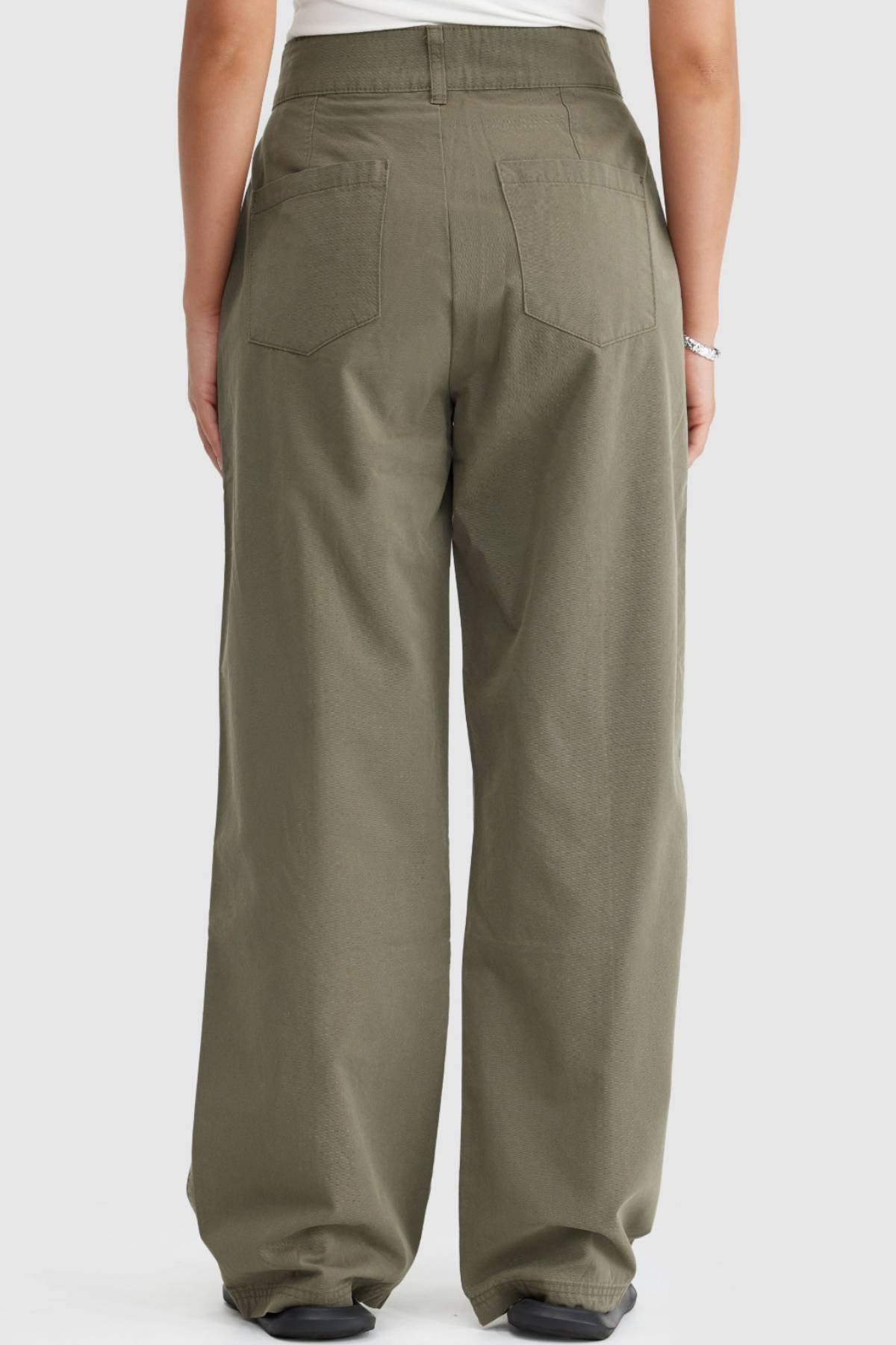 A person is shown from the waist down, back view, wearing loose-fitting olive green ORTC Avery Front Seam Pants with two back pockets and wide legs, paired with a white top. The pants are made from cotton tencel.