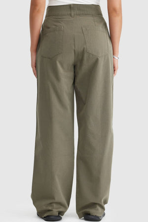 A person is shown from the waist down, back view, wearing loose-fitting olive green ORTC Avery Front Seam Pants with two back pockets and wide legs, paired with a white top. The pants are made from cotton tencel.