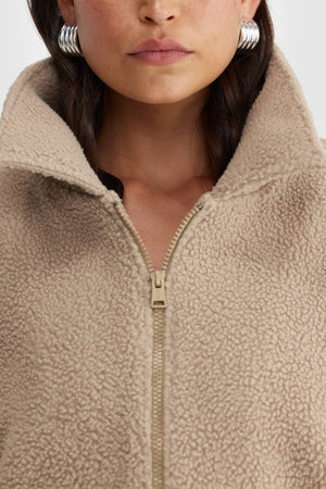A woman wears the ORTC Georgia Sherpa Jacket, a beige, textured piece made from recycled polyester, zipped halfway with the collar up, paired with large silver hoop earrings.