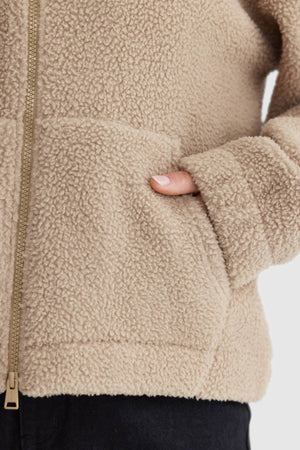 Someone wearing the ORTC Georgia Sherpa Jacket in beige, made from recycled polyester, with a hand in the fleece pocket. The jacket has a visible zipper and is paired with black pants.