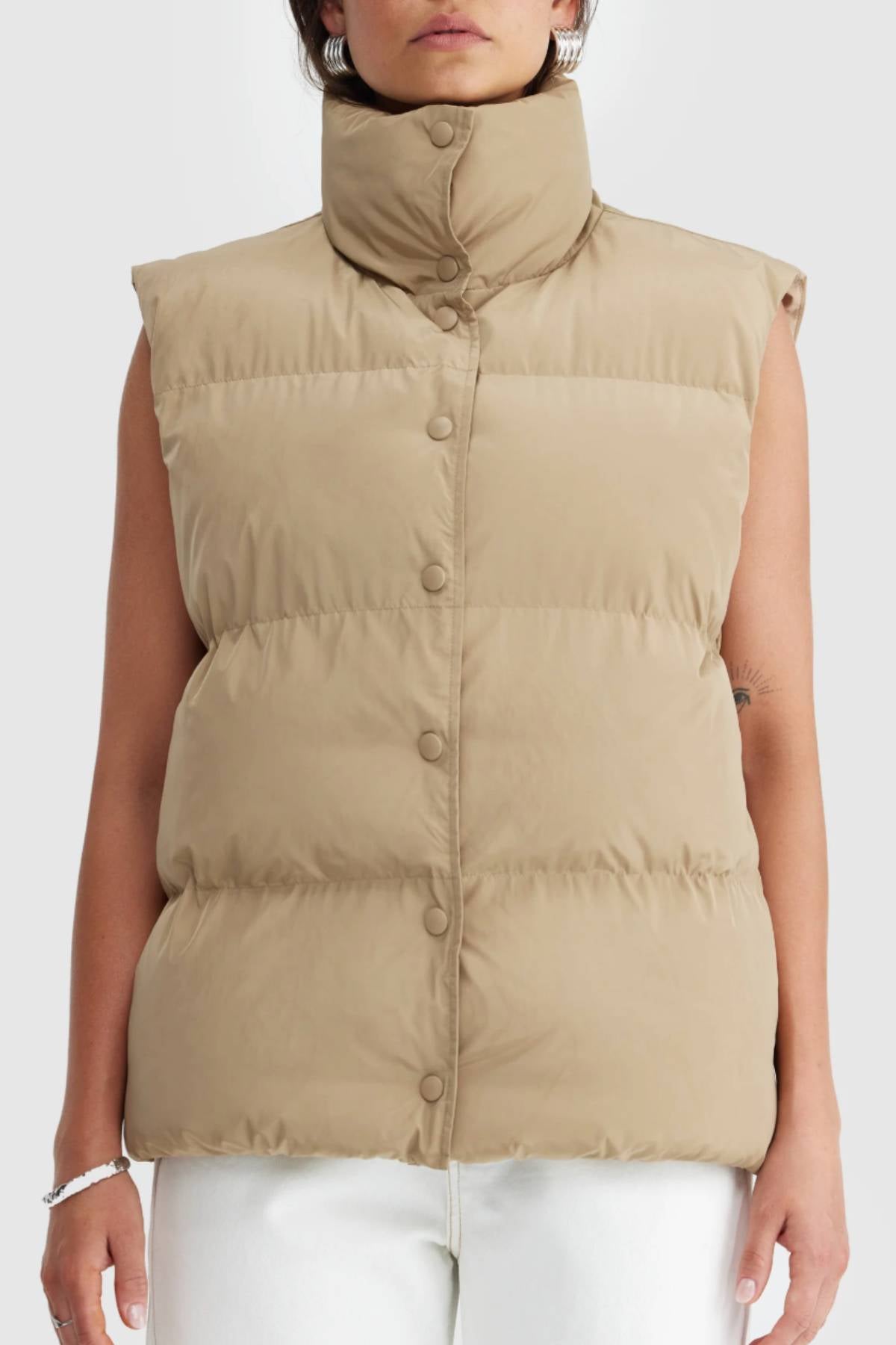 Someone wears the ORTC Montana Puffer Vest, a beige water-resistant, sleeveless vest with a high collar and snap buttons, styled with white pants. The vest is shown from shoulders to hips against a plain background.