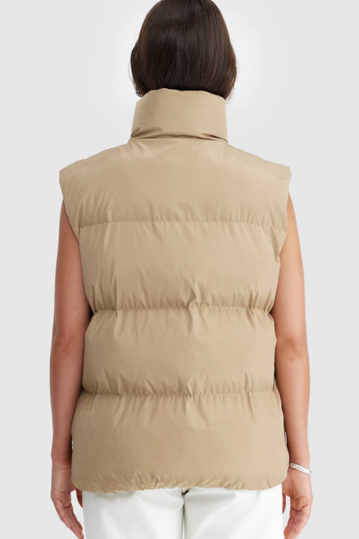 A person with shoulder-length brown hair is seen from behind wearing the ORTC Montana Puffer Vest in beige with a high collar and white pants, standing out against a plain, light-colored background.