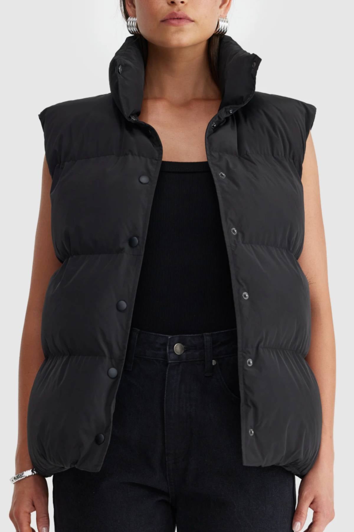 A woman wears the ORTC Montana Puffer Vest, black, insulated, buttoned over a black top and pants with the high collar turned up.