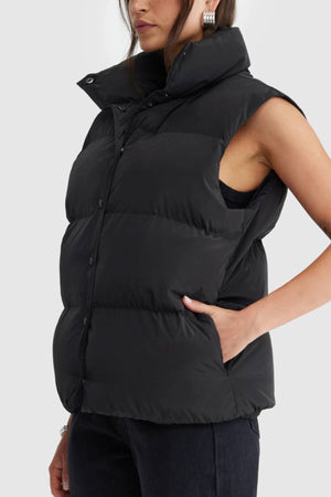 A woman wears the ORTC Montana Puffer Vest, a sleeveless, water-resistant vest with snap buttons, paired with dark pants. She stands with her left hand in her pocket, turned slightly to the side against a plain background.