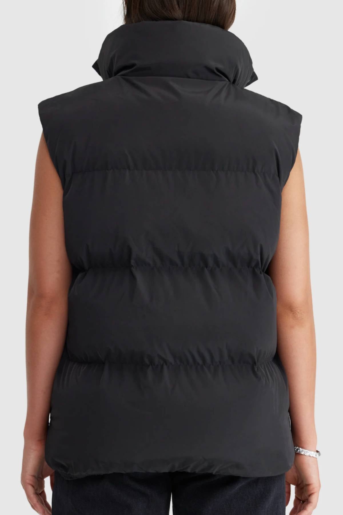 A person is seen from behind wearing an ORTC Montana Puffer Vest with a high collar, dark pants, and bare arms. The black insulated vest contrasts with the plain, light-colored background.