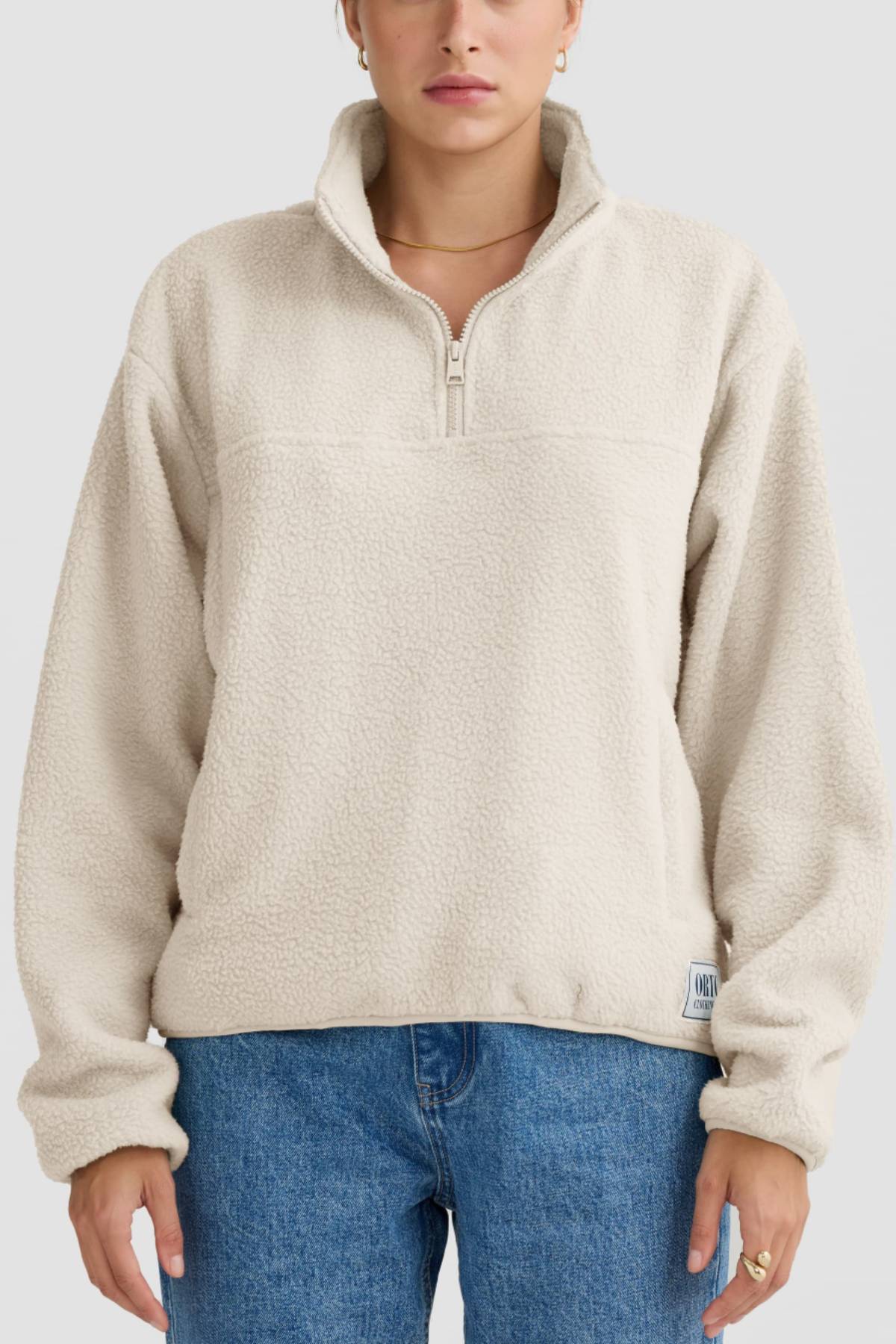 Someone wearing the ORTC Sherpa Fleece Quarter Zip in light beige, made from recycled polyester, with a high collar and relaxed fit, paired with blue jeans and standing against a plain light background.